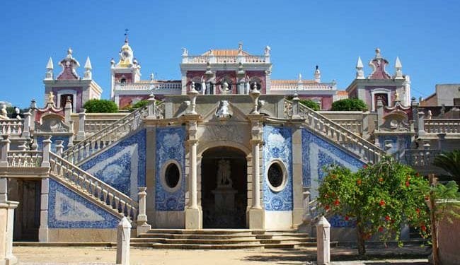 Un grande edificio storico con un'architettura ricercata e accenti di piastrelle blu è catturato sotto un cielo azzurro e limpido. Ampie scale in pietra con intricate ringhiere conducono ad un ingresso ad arco fiancheggiato da due finestre circolari. Una vegetazione lussureggiante circonda la scena, che ricorda il fascino che si prova quando si visitano gli aldeias dell'Algarve.