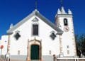 Una chiesa bianca ad Alte con una porta ad arco verde, cornici delle finestre decorate e un campanile con un orologio. La facciata presenta finestre simmetriche e decorative e una croce in cima al timpano. Visitare questo gioiello in Algarve sotto un cielo azzurro e limpido.