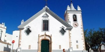 Una chiesa bianca ad Alte con una porta ad arco verde, cornici delle finestre decorate e un campanile con un orologio. La facciata presenta finestre simmetriche e decorative e una croce in cima al timpano. Visitare questo gioiello in Algarve sotto un cielo azzurro e limpido.