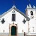 Una chiesa bianca ad Alte con una porta ad arco verde, cornici delle finestre decorate e un campanile con un orologio. La facciata presenta finestre simmetriche e decorative e una croce in cima al timpano. Visitare questo gioiello in Algarve sotto un cielo azzurro e limpido.