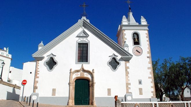 Una chiesa bianca ad Alte con una porta ad arco verde, cornici delle finestre decorate e un campanile con un orologio. La facciata presenta finestre simmetriche e decorative e una croce in cima al timpano. Visitare questo gioiello in Algarve sotto un cielo azzurro e limpido.