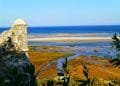 Una vista panoramica della costa con una storica torre di avvistamento in pietra sulla sinistra, che si affaccia su una spiaggia sabbiosa e un mare blu. Il primo piano presenta una vegetazione lussureggiante e zone umide, mentre l'orizzonte mostra un cielo azzurro e limpido che incontra l'oceano: una scena idilliaca che ti invita a visitare l'Algarve Cacela Velha.