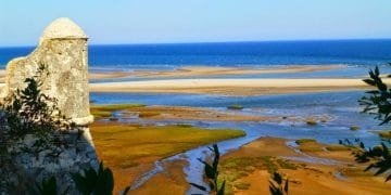 Una vista panoramica della costa con una storica torre di avvistamento in pietra sulla sinistra, che si affaccia su una spiaggia sabbiosa e un mare blu. Il primo piano presenta una vegetazione lussureggiante e zone umide, mentre l'orizzonte mostra un cielo azzurro e limpido che incontra l'oceano: una scena idilliaca che ti invita a visitare l'Algarve Cacela Velha.