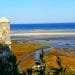 Una vista panoramica della costa con una storica torre di avvistamento in pietra sulla sinistra, che si affaccia su una spiaggia sabbiosa e un mare blu. Il primo piano presenta una vegetazione lussureggiante e zone umide, mentre l'orizzonte mostra un cielo azzurro e limpido che incontra l'oceano: una scena idilliaca che ti invita a visitare l'Algarve Cacela Velha.