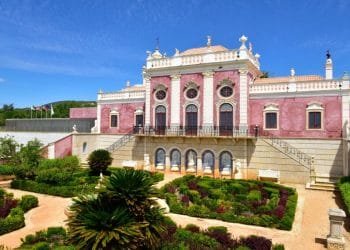 L'immagine mostra un grande ed elegante edificio simile a un castello con una facciata rosa e dettagli architettonici decorati. Di fronte, un giardino ben curato con siepi e sentieri ben tagliati ne esalta la grandiosità. Il cielo azzurro brillante si aggiunge alla vista panoramica, rendendolo una tappa obbligata per chiunque abbia intenzione di visitare Estói in Algarve.