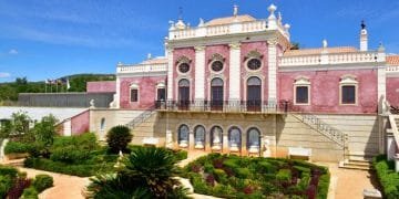 L'immagine mostra un grande ed elegante edificio simile a un castello con una facciata rosa e dettagli architettonici decorati. Di fronte, un giardino ben curato con siepi e sentieri ben tagliati ne esalta la grandiosità. Il cielo azzurro brillante si aggiunge alla vista panoramica, rendendolo una tappa obbligata per chiunque abbia intenzione di visitare Estói in Algarve.