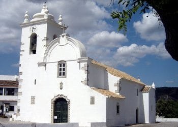 Una storica chiesa bianca con campanile e ingresso ad arco, sullo sfondo di un cielo azzurro e nuvole sparse. L'edificio presenta un'architettura tradizionale con tetto in tegole e piccole finestre. Un ramo di un albero con foglie verdi incornicia il lato destro dell'immagine, invitando a visitare l'Algarve Querença.