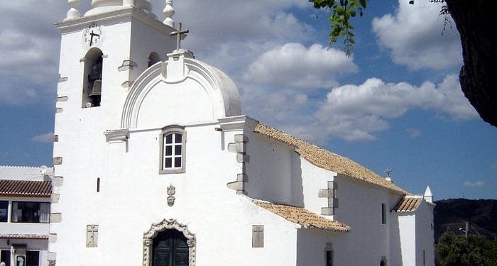 Una storica chiesa bianca con campanile e ingresso ad arco, sullo sfondo di un cielo azzurro e nuvole sparse. L'edificio presenta un'architettura tradizionale con tetto in tegole e piccole finestre. Un ramo di un albero con foglie verdi incornicia il lato destro dell'immagine, invitando a visitare l'Algarve Querença.