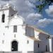 Una storica chiesa bianca con campanile e ingresso ad arco, sullo sfondo di un cielo azzurro e nuvole sparse. L'edificio presenta un'architettura tradizionale con tetto in tegole e piccole finestre. Un ramo di un albero con foglie verdi incornicia il lato destro dell'immagine, invitando a visitare l'Algarve Querença.