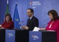 Tre persone stanno dietro i podi durante una conferenza stampa. La donna a sinistra è vestita di rosso, l'uomo al centro indossa un abito e anche la donna a destra è vestita di rosso e tiene in mano delle carte. Sullo sfondo sono raffigurate le bandiere europee e portoghesi con il testo "República Portuguesa Finanças", che indica alimenti essenziali IVA 0%.