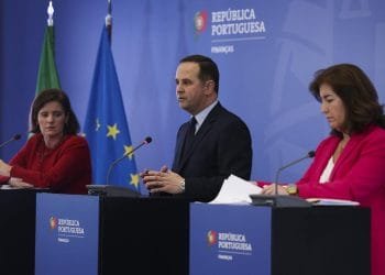 Tre persone stanno dietro i podi durante una conferenza stampa. La donna a sinistra è vestita di rosso, l'uomo al centro indossa un abito e anche la donna a destra è vestita di rosso e tiene in mano delle carte. Sullo sfondo sono raffigurate le bandiere europee e portoghesi con il testo "República Portuguesa Finanças", che indica alimenti essenziali IVA 0%.