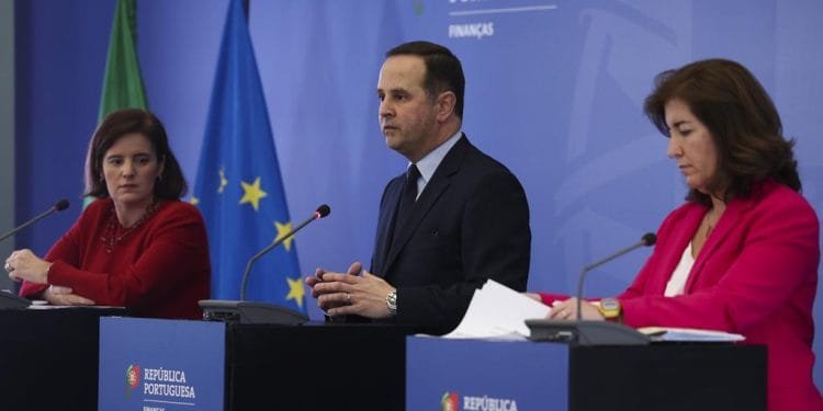Tre persone stanno dietro i podi durante una conferenza stampa. La donna a sinistra è vestita di rosso, l'uomo al centro indossa un abito e anche la donna a destra è vestita di rosso e tiene in mano delle carte. Sullo sfondo sono raffigurate le bandiere europee e portoghesi con il testo "República Portuguesa Finanças", che indica alimenti essenziali IVA 0%.