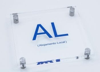 Un cartello in acrilico trasparente con supporti angolari metallici mostra il testo "AL" in grandi lettere blu e "(Imposta Straordinaria Alojamento Local)" in stampa blu più piccola sotto di esso, indicando una sistemazione locale. Lo sfondo è bianco.