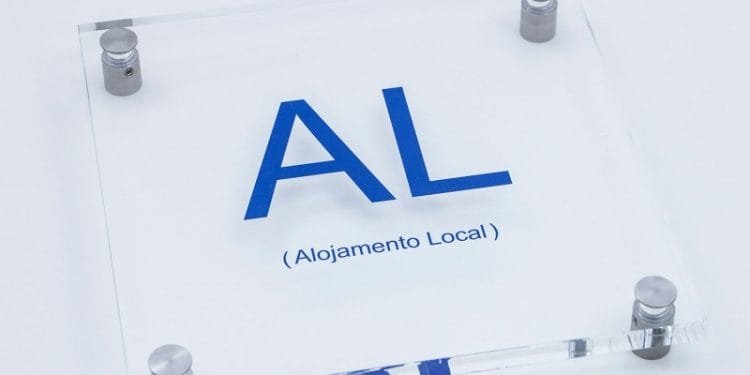 Un cartello in acrilico trasparente con supporti angolari metallici mostra il testo "AL" in grandi lettere blu e "(Imposta Straordinaria Alojamento Local)" in stampa blu più piccola sotto di esso, indicando una sistemazione locale. Lo sfondo è bianco.
