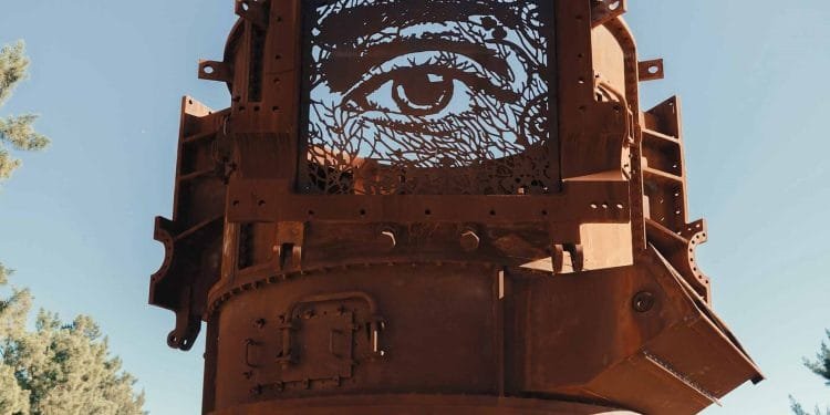 Una scultura in metallo arrugginito del famoso artista portoghese Vhils presenta un grande occhio dettagliato su un pannello quadrato. L'occhio ritagliato in modo intricato consente alla luce di passare attraverso, proiettando ombre dinamiche. Questo pezzo accattivante si trova all'aperto contro un cielo blu con alberi sullo sfondo.