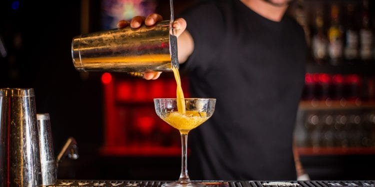 Un barista in camicia nera versa con cura un cocktail da uno shaker in un bicchiere a stelo. L'allestimento del bar, che ricorda un locale notturno di Albufeira, comprende varie bottiglie e uno sfondo poco illuminato con luci rosse e gialle, creando un'atmosfera calda e invitante, una scena degna di qualsiasi evento bartending della Gara internazionale.