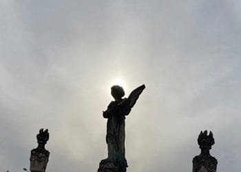 La sagoma di una statua di angelo con le ali ripiegate, in piedi di fronte al sole, creando un effetto alone attorno alla testa. Altre due statue più piccole lo fiancheggiano su entrambi i lati, tutte contro un cielo nuvoloso, evocando temi di mortalità in Portogallo.