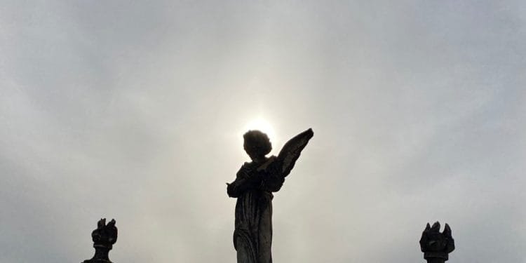 La sagoma di una statua di angelo con le ali ripiegate, in piedi di fronte al sole, creando un effetto alone attorno alla testa. Altre due statue più piccole lo fiancheggiano su entrambi i lati, tutte contro un cielo nuvoloso, evocando temi di mortalità in Portogallo.