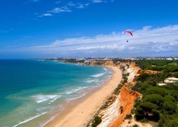 Una splendida costa in Algarve con spiagge di sabbia dorata incontra onde turchesi. Scogliere rosse e arancioni si innalzano sopra la riva, punteggiate di fogliame verde. Un parapendio con un paracadute rosso e bianco vola nel cielo azzurro e limpido, aggiungendo un senso di avventura a questa pittoresca spiaggia.