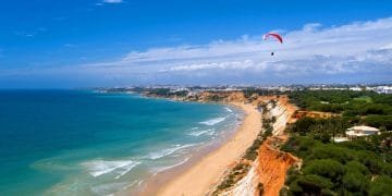 Una splendida costa in Algarve con spiagge di sabbia dorata incontra onde turchesi. Scogliere rosse e arancioni si innalzano sopra la riva, punteggiate di fogliame verde. Un parapendio con un paracadute rosso e bianco vola nel cielo azzurro e limpido, aggiungendo un senso di avventura a questa pittoresca spiaggia.