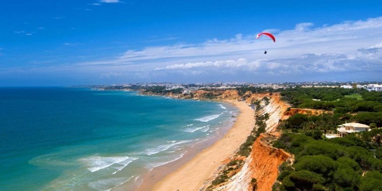 Una splendida costa in Algarve con spiagge di sabbia dorata incontra onde turchesi. Scogliere rosse e arancioni si innalzano sopra la riva, punteggiate di fogliame verde. Un parapendio con un paracadute rosso e bianco vola nel cielo azzurro e limpido, aggiungendo un senso di avventura a questa pittoresca spiaggia.