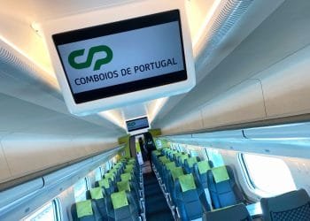 Interno di una moderna cabina ferroviaria con file di sedili blu e verdi ben organizzati. In alto, un display digitale mostra "CP" e "Comboios de Portugal", che indicano l'operatore ferroviario. Grandi finestre fiancheggiano le pareti, consentendo alla luce naturale di entrare nella cabina pulita e ben illuminata: davvero utile e positivo.