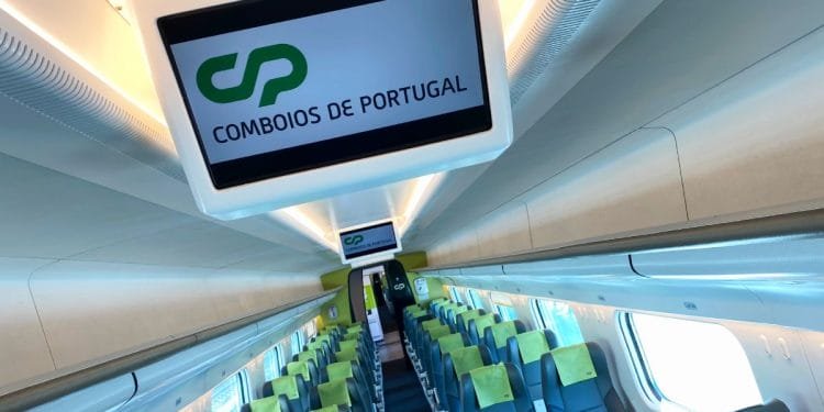 Interno di una moderna cabina ferroviaria con file di sedili blu e verdi ben organizzati. In alto, un display digitale mostra "CP" e "Comboios de Portugal", che indicano l'operatore ferroviario. Grandi finestre fiancheggiano le pareti, consentendo alla luce naturale di entrare nella cabina pulita e ben illuminata: davvero utile e positivo.