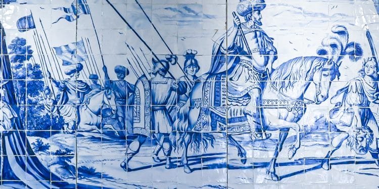 Un dettagliato murale in piastrelle bianche e blu raffigura una scena storica con soldati a cavallo e a piedi, che portano bandiere e stendardi. La figura centrale è un condottiero in armatura a cavallo, circondato da truppe e ombreggiato da alberi sullo sfondo, che celebra la Giornata internazionale monumenti siti.