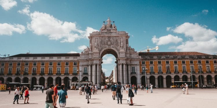 Le persone camminano attraverso una grande piazza aperta con un grande arco al centro, sotto un cielo limpido e azzurro. L'arco, ornato di sculture, è fiancheggiato da edifici dalle facciate gialle. La scena è luminosa e vivace, ricorda le vivaci piazze di città come Lisbona e Porto.