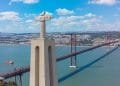 Una grande statua di Cristo con le braccia tese si erge su un alto piedistallo di cemento che si affaccia su un ampio fiume. Sullo sfondo, un ponte sospeso rosso attraversa il fiume, incorniciato dal paesaggio urbano e dalle colline di Lisbona, la città più cara d'Europa, sotto un cielo azzurro brillante con nuvole sparse.
