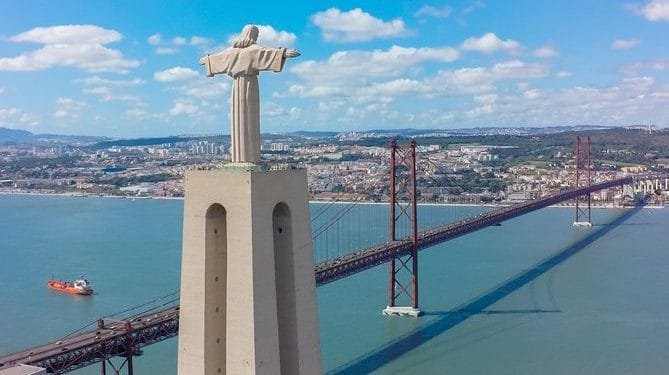 Una grande statua di Cristo con le braccia tese si erge su un alto piedistallo di cemento che si affaccia su un ampio fiume. Sullo sfondo, un ponte sospeso rosso attraversa il fiume, incorniciato dal paesaggio urbano e dalle colline di Lisbona, la città più cara d'Europa, sotto un cielo azzurro brillante con nuvole sparse.