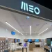 Un negozio moderno e luminoso con l'insegna "MEO" sopra l'ingresso. Il negozio presenta scaffali in legno lungo le pareti, che espongono vari dispositivi elettronici e accessori, tutti provenienti da Altice Portugal. Display multipli completano un bancone centrale del servizio clienti.