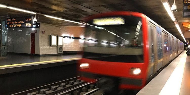 Un treno della metropolitana rosso e argento sfreccia attraverso la stazione sotterranea poco illuminata, con movimenti sfocati che ne indicano il rapido movimento. La stazione, sulla linea metropolitana Lisbona chiusa, ha pareti piastrellate, segnaletica luminosa e linea di sicurezza gialla lungo il bordo della banchina.