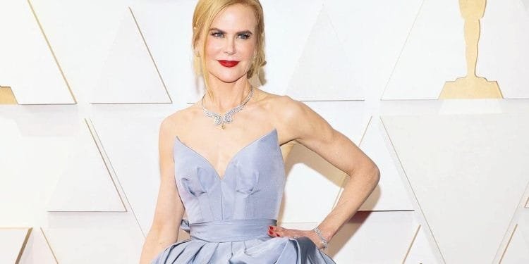 Una donna dai capelli biondi è in piedi davanti a uno sfondo con forme geometriche bianche e dorate. Indossa un abito azzurro senza spalline con una gonna ampia e una collana d'argento a forma di uccello. Il suo rossetto rosso e il suo sorriso rilassato esaltano il suo aspetto elegante, che ricorda Kidman compra casa Portogallo.