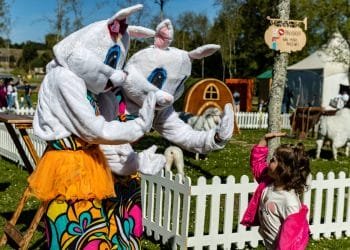 Due persone in allegri costumi da coniglio danno il cinque a una giovane ragazza con una giacca rosa durante un evento all'aperto che celebra la Pasqua Portogallo. Si trovano in un'area recintata da una staccionata bianca, con altri personaggi in costume e recinti di piccoli animali visibili sullo sfondo.
