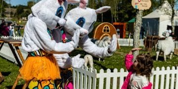 Due persone in allegri costumi da coniglio danno il cinque a una giovane ragazza con una giacca rosa durante un evento all'aperto che celebra la Pasqua Portogallo. Si trovano in un'area recintata da una staccionata bianca, con altri personaggi in costume e recinti di piccoli animali visibili sullo sfondo.