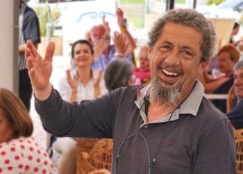 Un uomo barbuto con i capelli grigi sorride e gesticola con la mano, circondato da una folla di persone, alcune delle quali applaudono e sorridono. L'ambientazione sembra essere informale, forse durante un incontro sociale o una celebrazione. Sembra che Raffaele Salomone si stia godendo appieno il momento.