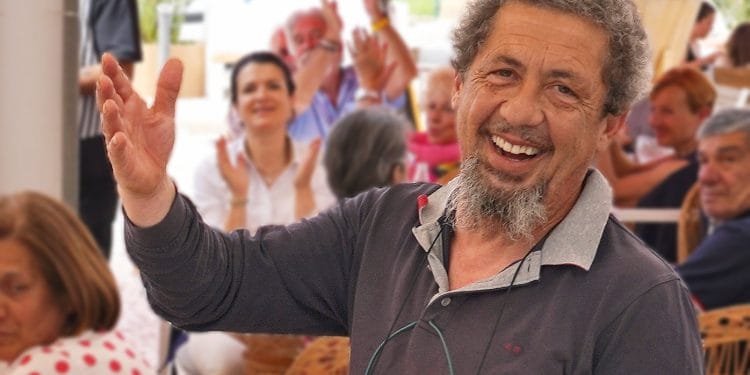 Un uomo barbuto con i capelli grigi sorride e gesticola con la mano, circondato da una folla di persone, alcune delle quali applaudono e sorridono. L'ambientazione sembra essere informale, forse durante un incontro sociale o una celebrazione. Sembra che Raffaele Salomone si stia godendo appieno il momento.