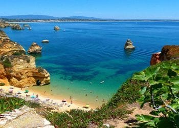 Una vista panoramica di una spiaggia soleggiata con acque turchesi, formazioni rocciose e una costa lontana. Le persone si rilassano sotto gli ombrelloni sulla riva sabbiosa. In primo piano sono visibili piante verdi rigogliose e un sentiero, che si affaccia su una delle migliori spiagge che Lisbona ha da offrire.