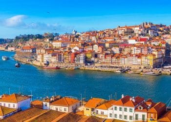 Una vivace scena di Porto, dove Tassa soggiorno in Portogallo è di 1,5 euro. Si vedono edifici colorati accatastati su una collina. In primo piano il fiume Douro con le barche e alcuni gabbiani che volano sopra. Il cielo è limpido e azzurro, esaltando la vista pittoresca.