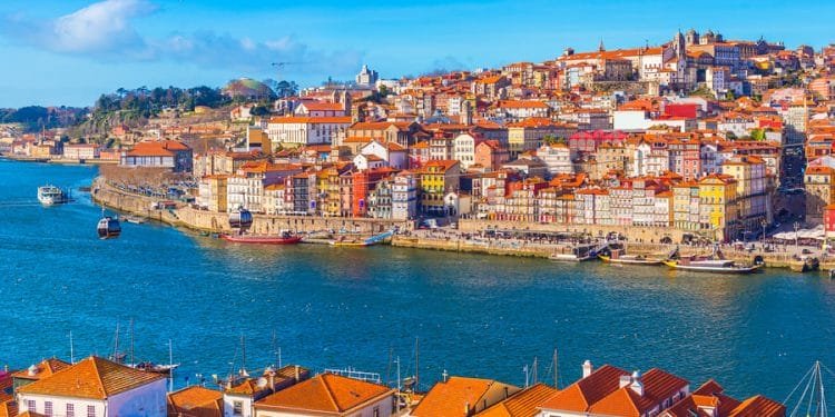 Una vivace scena di Porto, dove Tassa soggiorno in Portogallo è di 1,5 euro. Si vedono edifici colorati accatastati su una collina. In primo piano il fiume Douro con le barche e alcuni gabbiani che volano sopra. Il cielo è limpido e azzurro, esaltando la vista pittoresca.