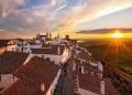 L'immagine mostra un pittoresco villaggio con case bianche e tetti di tegole rosse, allineato lungo una stretta strada acciottolata. Il sole sta tramontando all'orizzonte, proiettando una calda luce sul paesaggio di campi verdi e acque lontane. È un invito a visitare le aldeie dell'Alentejo e a sperimentarne il fascino.