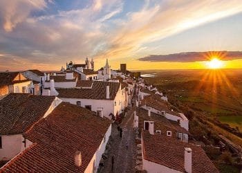 L'immagine mostra un pittoresco villaggio con case bianche e tetti di tegole rosse, allineato lungo una stretta strada acciottolata. Il sole sta tramontando all'orizzonte, proiettando una calda luce sul paesaggio di campi verdi e acque lontane. È un invito a visitare le aldeie dell'Alentejo e a sperimentarne il fascino.