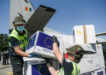 Due lavoratori in uniforme e cappello stanno scaricando scatole da un aereo, etichettate con la bandiera dell'Unione Europea e la scritta "rescEU". Le scatole contengono assistenza medica critica per catastrofi. Entrambi i lavoratori indossano maschere blu e la coda dell'aereo presenta una bandiera rossa, gialla e blu del Portogallo.