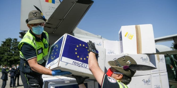 Due lavoratori in uniforme e cappello stanno scaricando scatole da un aereo, etichettate con la bandiera dell'Unione Europea e la scritta "rescEU". Le scatole contengono assistenza medica critica per catastrofi. Entrambi i lavoratori indossano maschere blu e la coda dell'aereo presenta una bandiera rossa, gialla e blu del Portogallo.