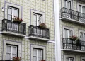 La facciata di un edificio presenta piastrelle a motivi geometrici e balconi con ringhiere decorate in ferro battuto. Diversi balconi sono adornati con vasi di fiori rossi, in contrasto con le piastrelle chiare. Le finestre sono incorniciate di bianco e hanno tende di pizzo, dando un senso di tranquillità mentre i residenti si godono i loro giorni a casa.