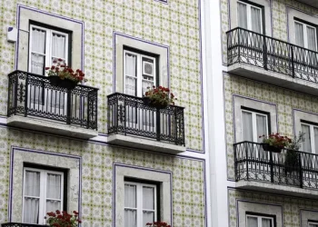La facciata di un edificio presenta piastrelle a motivi geometrici e balconi con ringhiere decorate in ferro battuto. Diversi balconi sono adornati con vasi di fiori rossi, in contrasto con le piastrelle chiare. Le finestre sono incorniciate di bianco e hanno tende di pizzo, dando un senso di tranquillità mentre i residenti si godono i loro giorni a casa.
