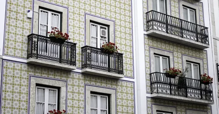 La facciata di un edificio presenta piastrelle a motivi geometrici e balconi con ringhiere decorate in ferro battuto. Diversi balconi sono adornati con vasi di fiori rossi, in contrasto con le piastrelle chiare. Le finestre sono incorniciate di bianco e hanno tende di pizzo, dando un senso di tranquillità mentre i residenti si godono i loro giorni a casa.