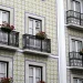 La facciata di un edificio presenta piastrelle a motivi geometrici e balconi con ringhiere decorate in ferro battuto. Diversi balconi sono adornati con vasi di fiori rossi, in contrasto con le piastrelle chiare. Le finestre sono incorniciate di bianco e hanno tende di pizzo, dando un senso di tranquillità mentre i residenti si godono i loro giorni a casa.
