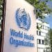 Un cartello con la scritta "Organizzazione Mondiale della Sanità" accanto all'emblema dell'OMS, raffigurante un bastone con un serpente e una mappa del mondo, è montato all'aperto con un edificio per uffici sfocato e alberi sullo sfondo, evidenziando gli sforzi dell'OMS contro la febbre dengue in Portogallo.